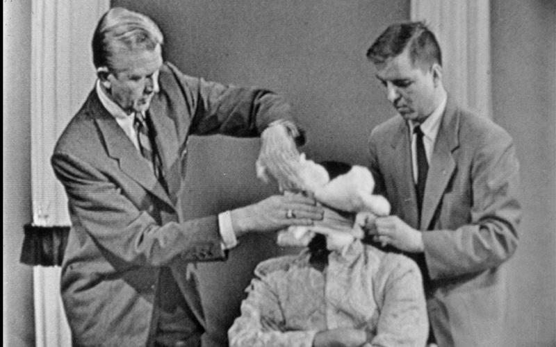 Two men in suits bandage a man's head to blindfold him